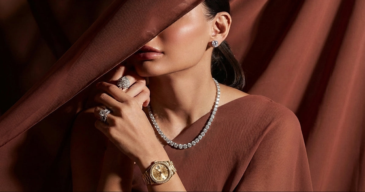 Woman wearing jewelry, including a necklace, earrings, and rings, with a brown curtain background.